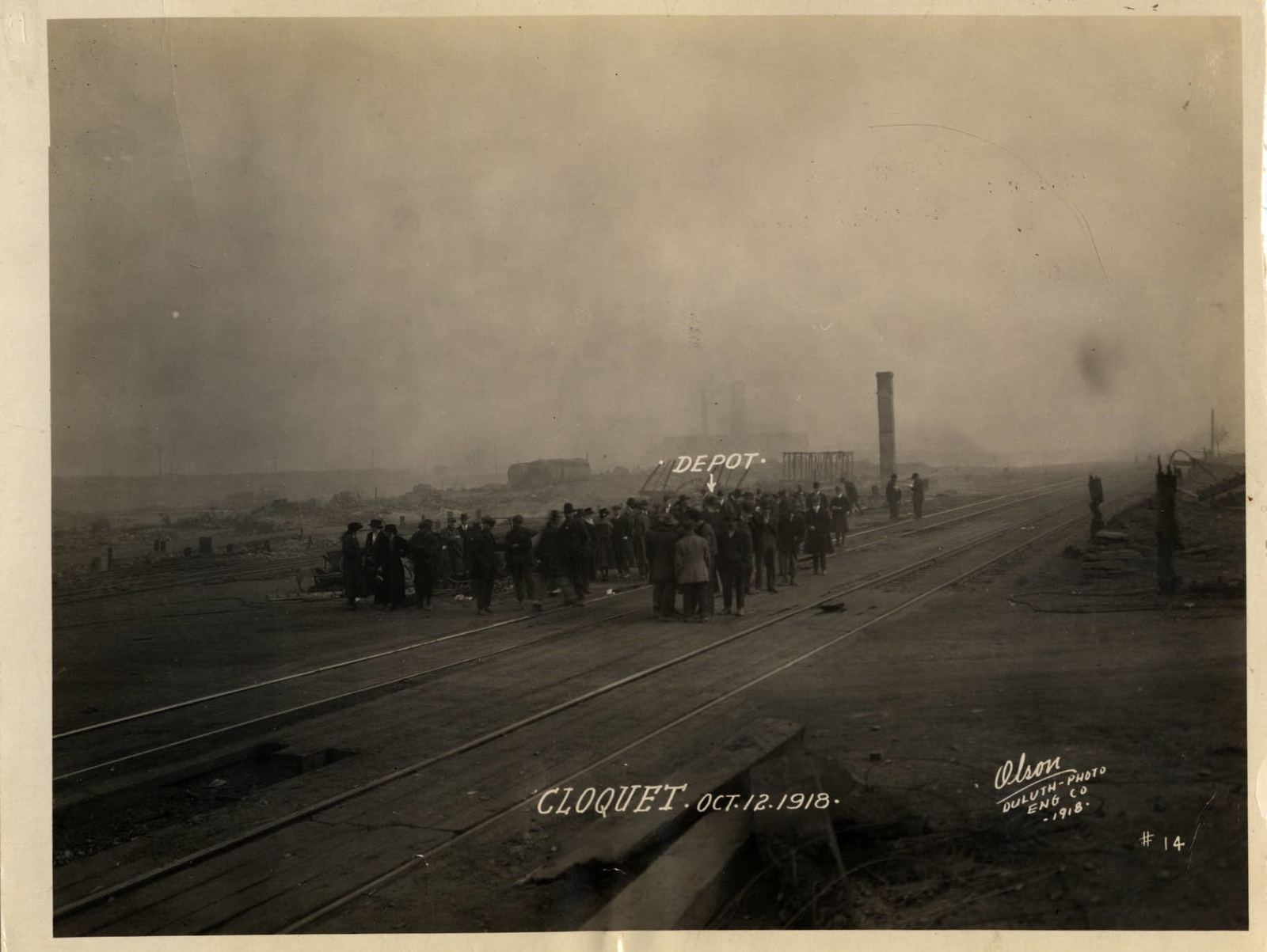 A crowd of men in coats and hats stands on the rail tracks where the Cloquet depot stood, with smoke still hanging over the town